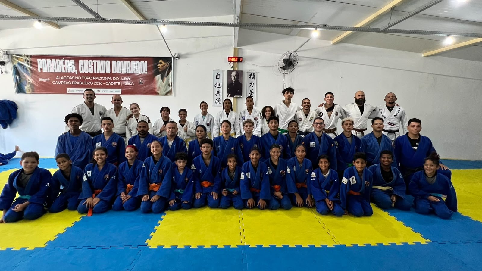 Grupo de judocas — crianças, jovens e adultos — reunido sobre um tatame azul e amarelo. Os atletas na frente estão sentados vestindo kimonos azuis; ao fundo, outros atletas e instrutores estão em pé usando kimonos brancos e azuis. Na parede posterior, há uma faixa vermelha com a frase "Parabéns, Gustavo Dourado! Alagoas no topo nacional do judô! Campeão Brasileiro 2026 - Cadete | -66kg"
